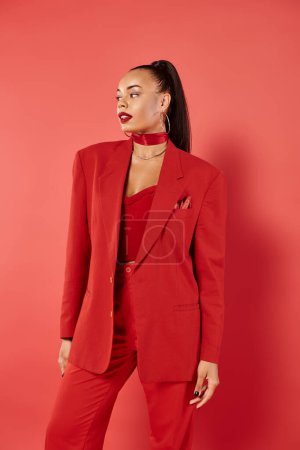 brunette african american woman with ponytail posing in suit and smiling on red background
