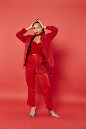 full length of brunette african american woman with ponytail posing in suit on red background