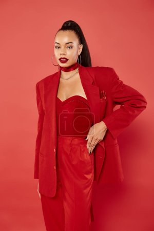 beautiful african american woman with ponytail posing in suit with hand on hip on red background
