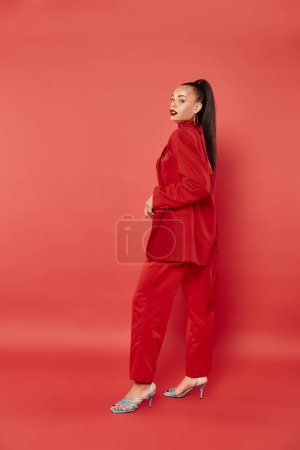 longitud completa de hermosa mujer afroamericana con cola de caballo posando en traje sobre fondo rojo