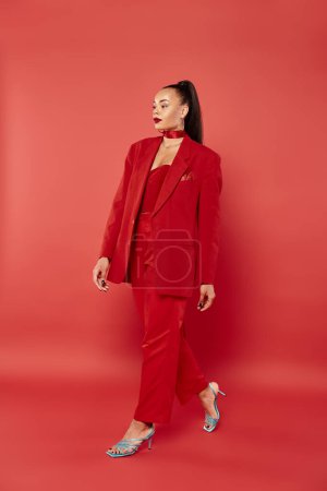 full length of beautiful african american woman with ponytail walking in suit on red background