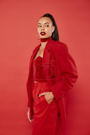 beautiful african american woman with ponytail posing in suit with hand in pocket on red background