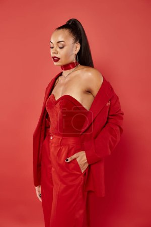 attractive african american woman with ponytail posing in suit with hand in pocket on red background