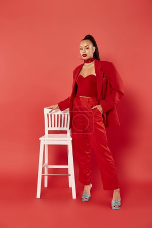 full length of attractive african american woman posing in red suit with hand in pocket near chair