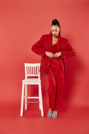 full length of attractive african american woman posing in vibrant suit near white chair on red
