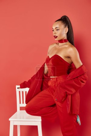beautiful african american woman posing in vibrant suit near white chair on red background