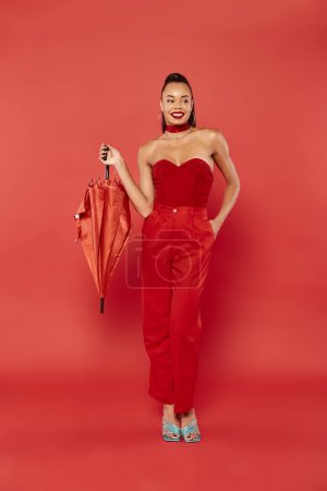 full length of happy african american model in red strapless top and pants posing with umbrella