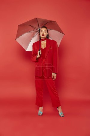 full length, brunette african american model in red blazer and pants standing under umbrella