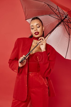 brunette african american woman in suit jacket and pants standing under umbrella on red backdrop
