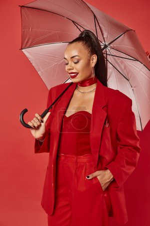 smiling african american woman in blazer and pants standing under umbrella on red backdrop