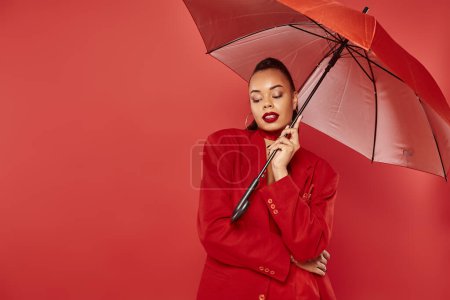 attractive young african american woman in blazer and pants standing under umbrella on red backdrop