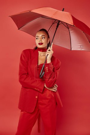 attractive young african american woman in jacket and pants standing under umbrella on red backdrop