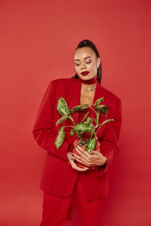 brunette young african american woman in red suit jacket and pants standing with potted green plant