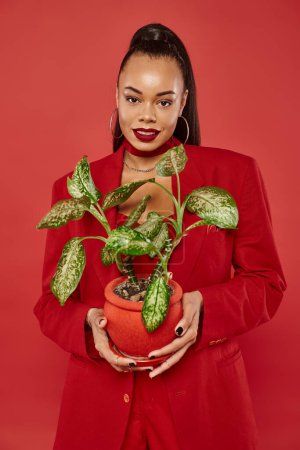 positive young african american woman in red suit jacket and pants standing with potted green plant