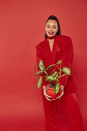 positive african american woman in red suit pants and jacket standing with potted green plant
