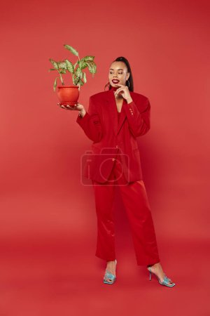 volle Länge, fröhliche afrikanisch-amerikanische Frau in roter formeller Kleidung mit grünen Topfpflanzen