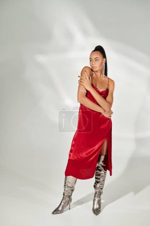 Foto de Longitud completa de morena mujer afroamericana en vestido rojo y botas de plata sobre fondo gris - Imagen libre de derechos