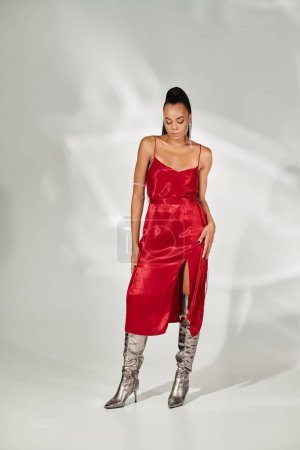 full length, african american woman in red midi dress and silver boots posing on grey backdrop