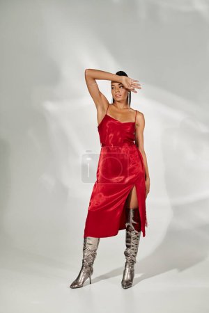 Foto de Mujer afroamericana en vestido midi rojo y botas plateadas posando sobre fondo gris espejado - Imagen libre de derechos