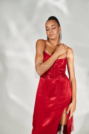 sensual african american woman in red dress looking away and posing on mirrored grey backdrop