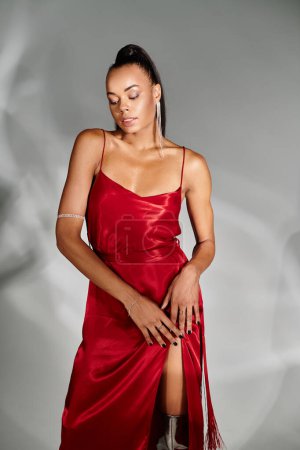 alluring african american woman in red dress looking down and posing on mirrored grey backdrop