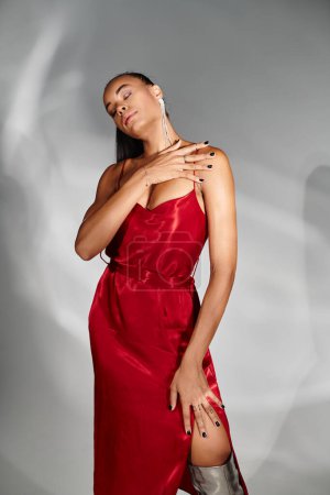 alluring african american woman in red dress posing with closed eyes on mirrored grey backdrop