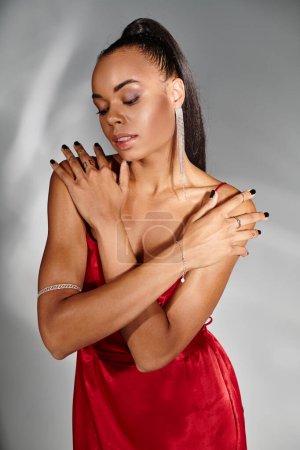 verführerische afrikanisch-amerikanische Frau im roten Kleid, die die Arme verschränkt, wenn sie vor verspiegeltem grauen Hintergrund posiert