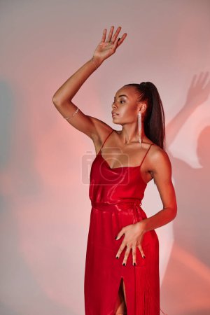 glamorous african american woman in red dress posing with raised hand on grey backdrop with lighting