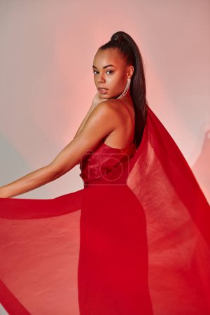 Foto de Encantadora mujer afroamericana en vestido rojo posando con chal sobre fondo gris con iluminación - Imagen libre de derechos