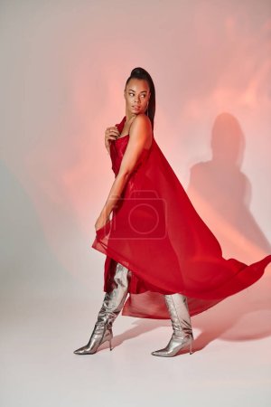 Foto de Mujer afroamericana en vestido rojo y chal caminando en botas plateadas sobre gris con iluminación - Imagen libre de derechos