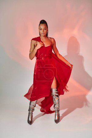 Afroamerikanerin in rotem Kleid mit Schal steht in silbernen Stiefeln auf grau mit Beleuchtung