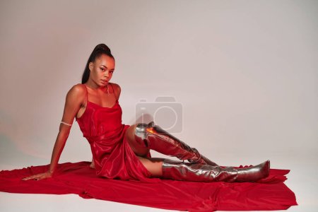 attraktive afrikanisch-amerikanische Frau in rotem Midikleid und silbernen Stiefeln sitzt auf grau mit Beleuchtung