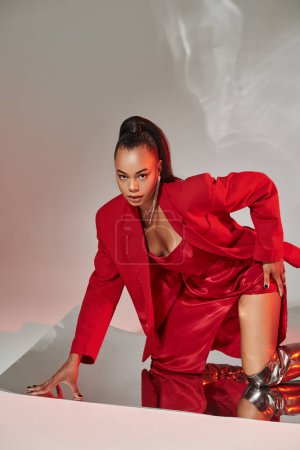 Foto de Joven modelo afroamericano en vestido rojo, chaqueta y botas plateadas posando sobre superficie espejada - Imagen libre de derechos