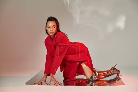 joven modelo afroamericano en blazer rojo, vestido y botas plateadas posando sobre superficie espejada