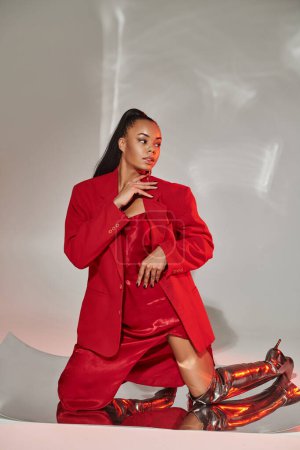 young african american woman in red blazer, dress and silver boots posing on mirrored surface