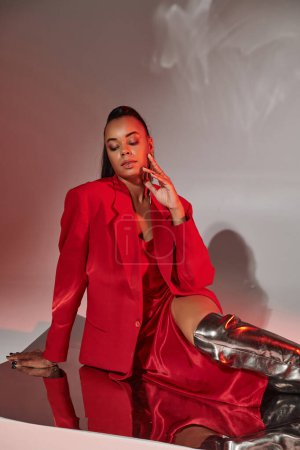 jolie femme afro-américaine en blazer rouge, robe et bottes argentées posant sur une surface miroir