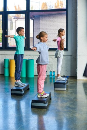 three adolescent girls and boy in sportswear exercising on fitness stepper with arms asides, sport