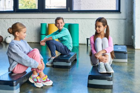two pretty girls and boy sitting on fitness steppers and smiling at each other, child sport