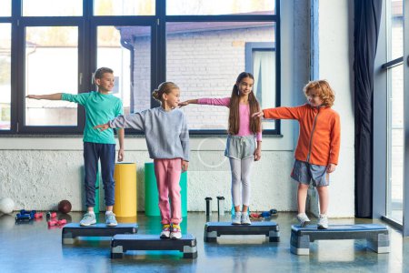 Foto de Cuatro pequeños niños lindos en ropa deportiva estirando los brazos de pie pasos de fitness, deporte infantil - Imagen libre de derechos