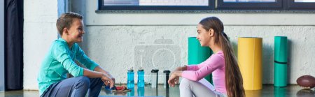 joyful boy and girl in sportswear sitting and smiling cheerfully at each other, child sport, banner