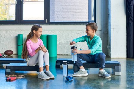 fröhliche vorpubertäre Kinder in Sportbekleidung sitzen auf Fitness-Steppschuhen mit Wasserflaschen, Sport