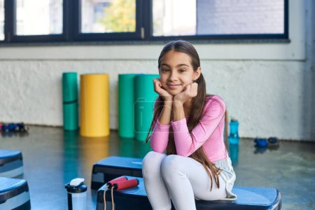 joyful girl sitting on fitness stepper with jump rope smiling at camera, hands under chin, sport