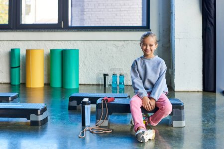 fröhliches blondes kleines Mädchen sitzt auf Fitness-Stepper in Sportbekleidung lächelnd in die Kamera, Kindersport
