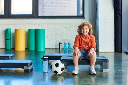 joyful red haired boy sitting on fitness stepper next to soccer ball and water bottle, child sport