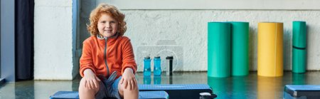 Photo for Happy red haired boy sitting on fitness stepper smiling cheerfully at camera, child sport, banner - Royalty Free Image