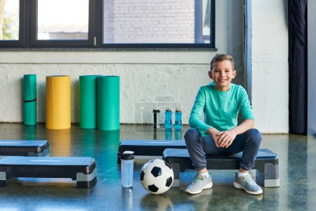 cheerful preadolescent boy on fitness stepper next to soccer ball and water bottles, child sport