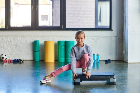 Photo for Little pretty girl in sportswear stretching leg on fitness stepper looking at camera, child sport - Royalty Free Image