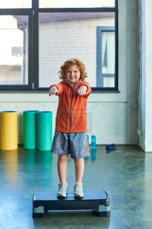 vertical shot of jolly little boy stretching arms on fitness stepper, smiling at camera, child sport