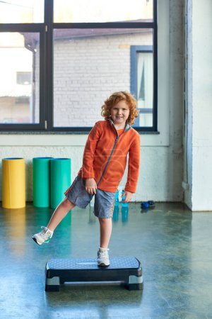 vertikale Aufnahme eines rothaarigen, fröhlichen Jungen, der auf einem Bein auf einem Fitness-Stepper in der Turnhalle steht, Kindersport