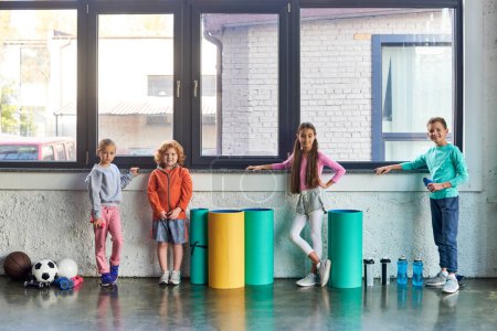 four preadolescent cute boys and girls in sportswear posing by window in gym, child sport